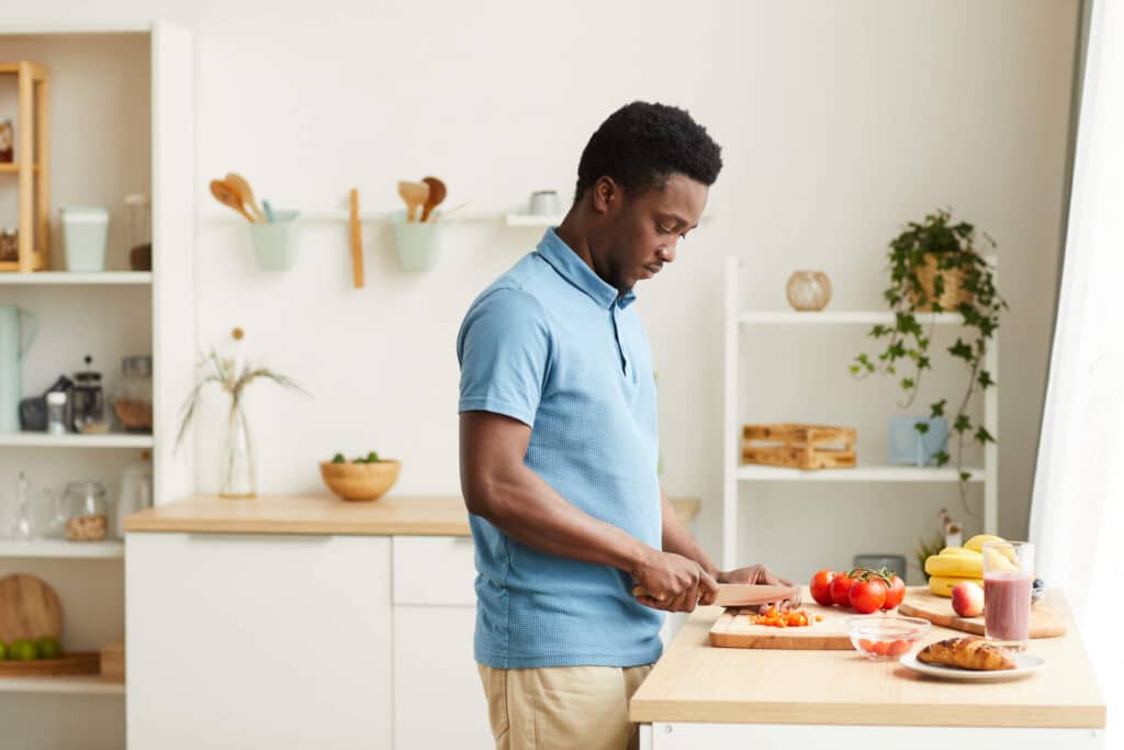 A man wearing blue shirt seriously slicing vegetables on a cutting board while standing in the middle of the kitchen
