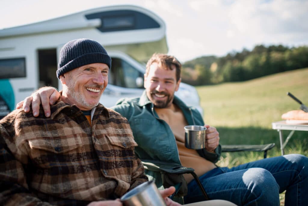 Two smiling men enjoying camping, sitting outside a camper van while sipping coffee