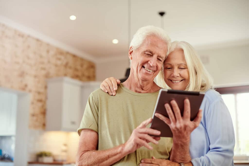 Happy elderly couple standing in the middle of living room while talking to a therapist through their ipad