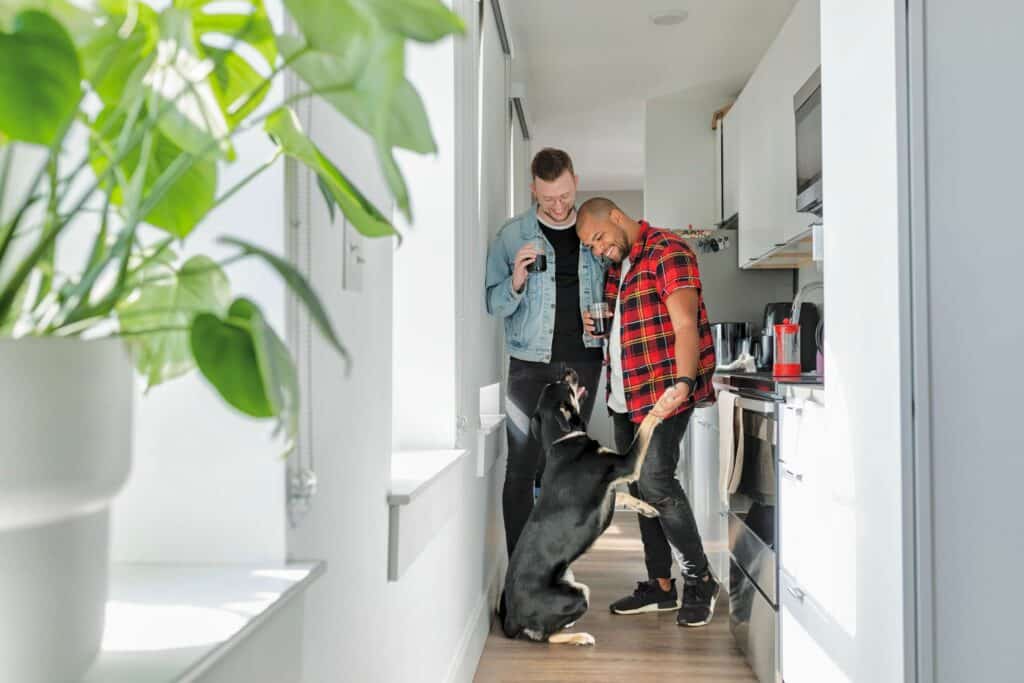 Two smiling men standing in the middle of the kitchen looking at a dog sitting on the floor