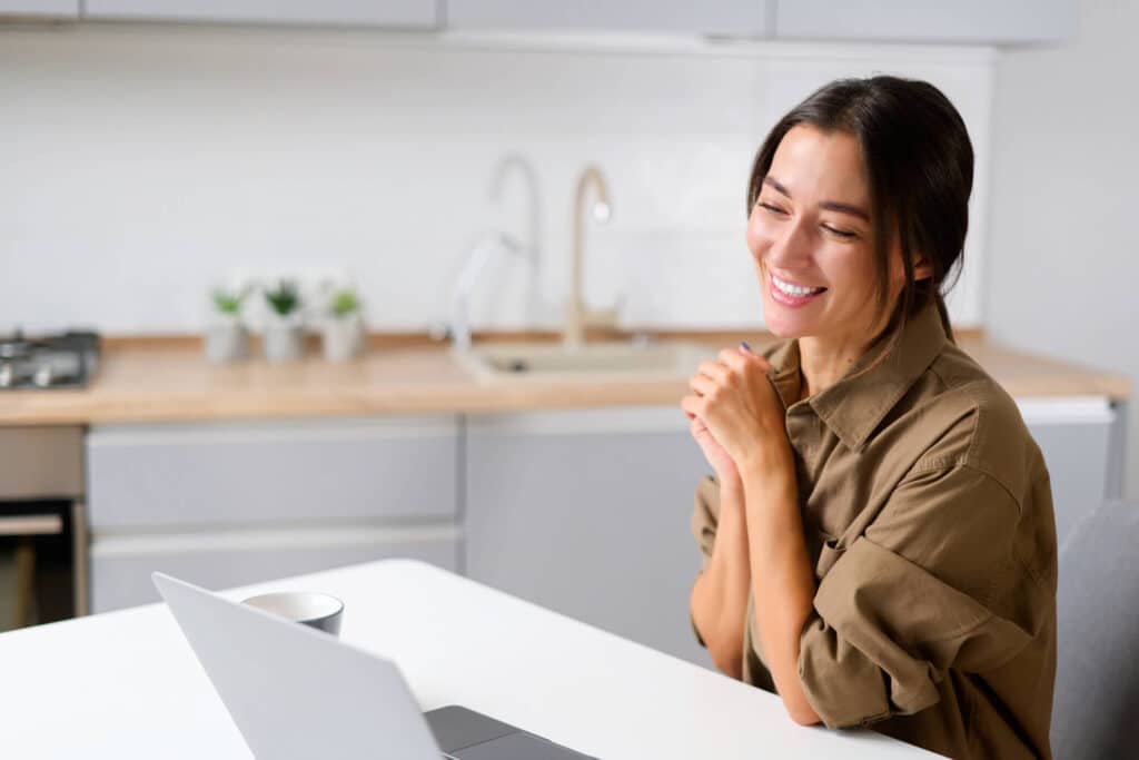 A lady wearing brown long sleeves smiling in front of the computer while sitting in the middle of the kitchen