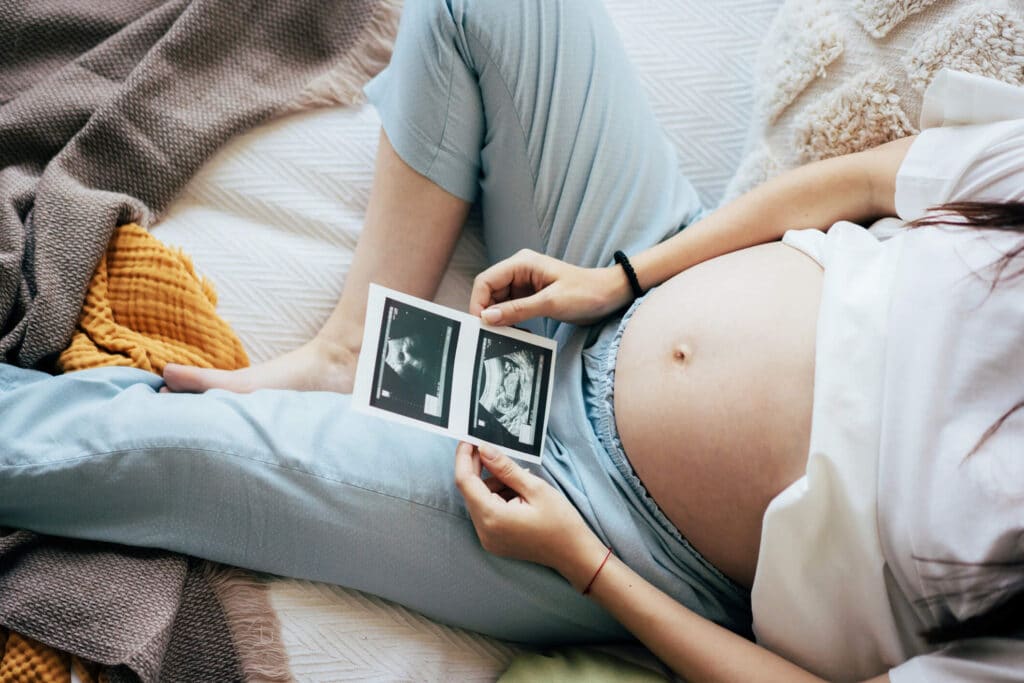 A pregnant woman showing her baby bump while sitting on a couch holding a sonogram