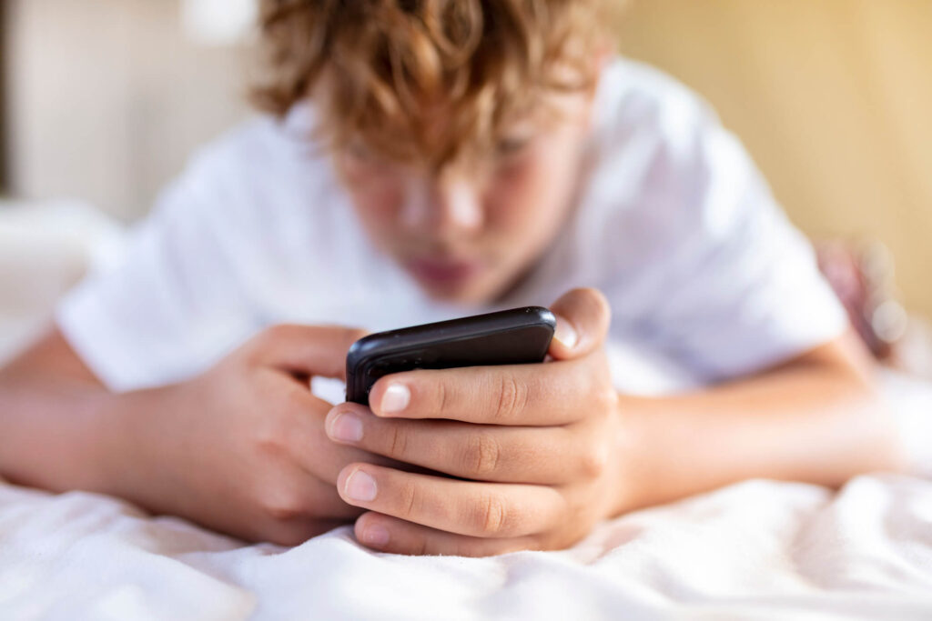 A young boy wearing white shirt lying on a bed while engrossed with his cell phone