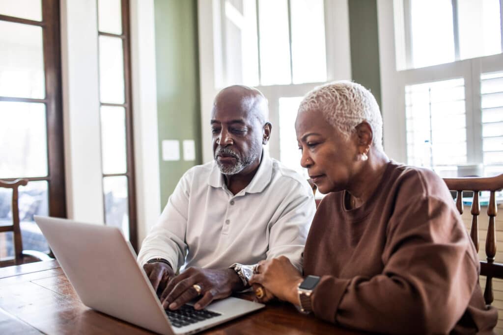An elderly man and woman sitting down in front of a laptop with neutral expressions, reading about therapy skills