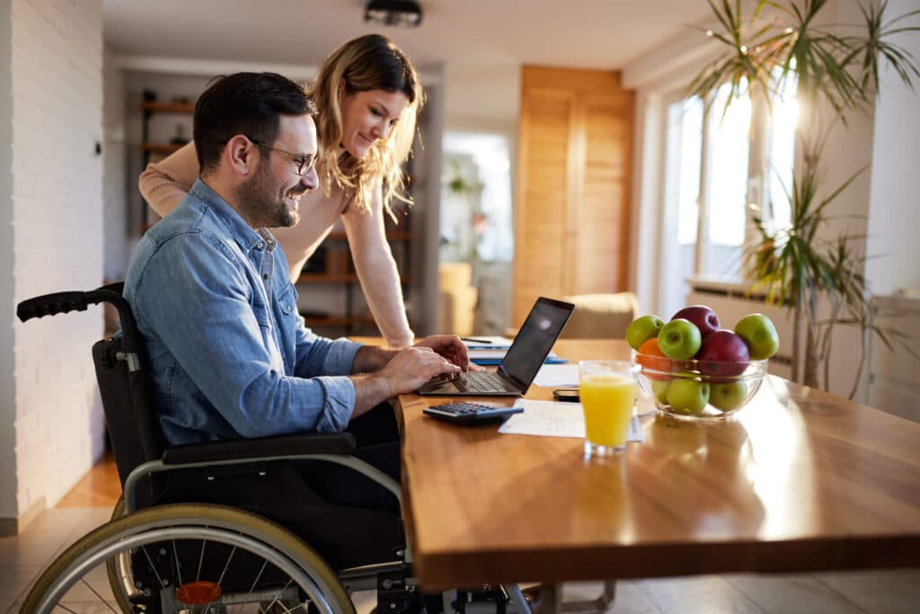 A man in a wheelchair and a woman standing next to him using a laptop and engaged in a productive interaction