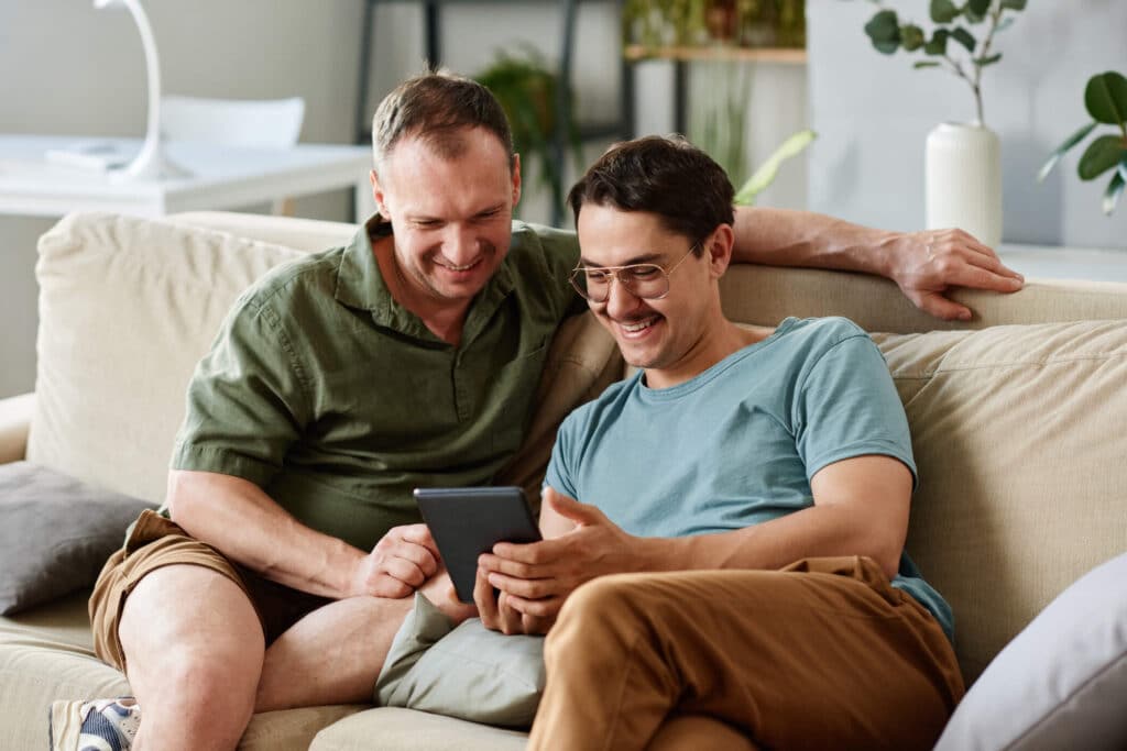 A pair of smiling men lounging on a couch while using a tablet