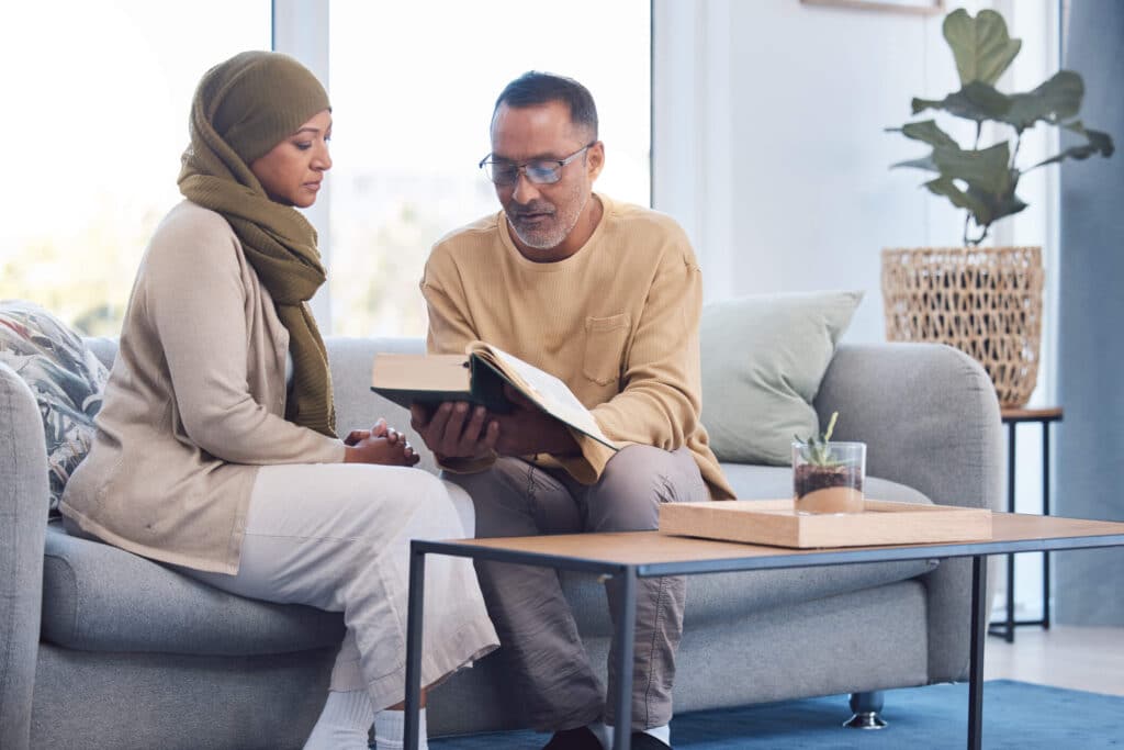 A woman wearing hijab and a man holding a bible both sitting on a couch sharing peaceful devotion