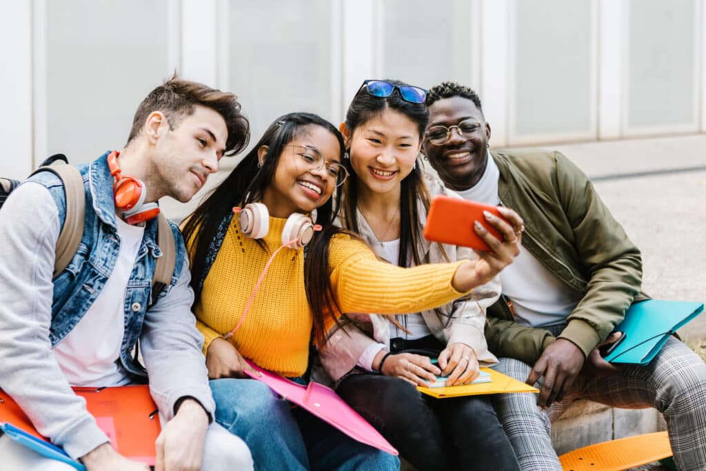 Group of happy young students posing in front of a camera capturing a selfie while sitting on a bench