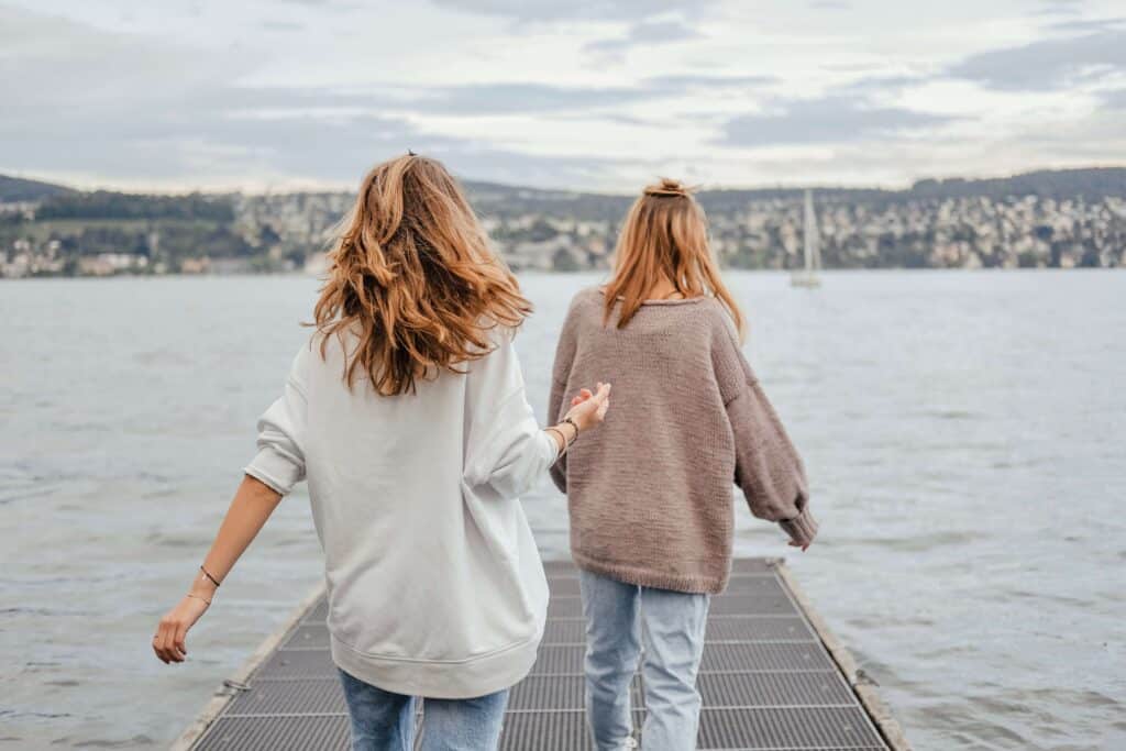 Two women wearing sweater walking towards the end of the dock with boat and mountain in the backdrop