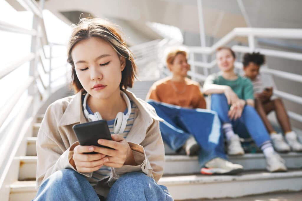 A sad young lady using her phone while sitting on steps with a group of kids sitting behind her