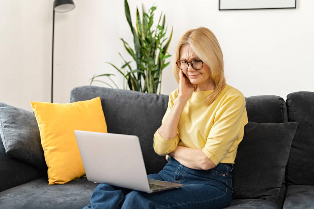 A woman wearing a yellow blouse sitting comfortably on a black couch while focused on her laptop