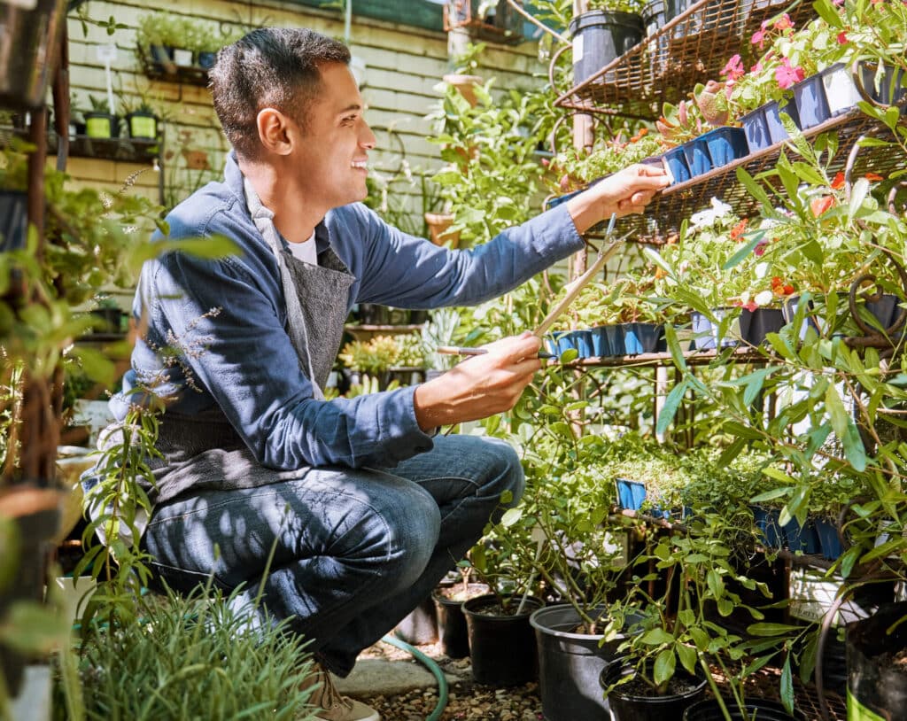 A man wearing a black apron sitting down in the middle of a green house smiling while checking the plants