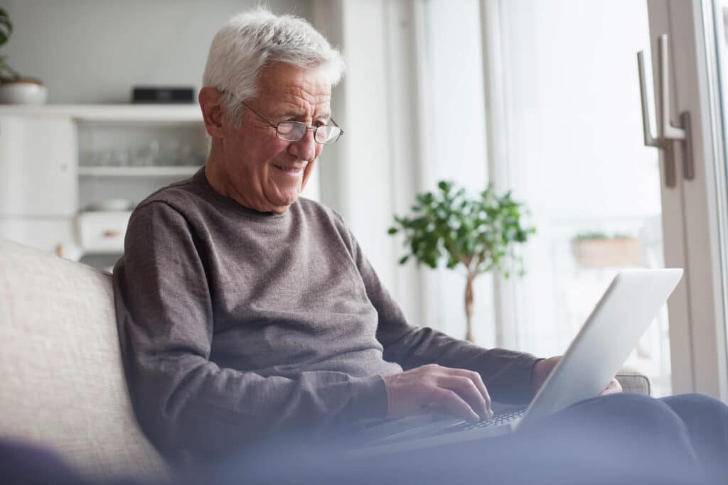 An elderly man sitting on a couch smiling in front of his laptop