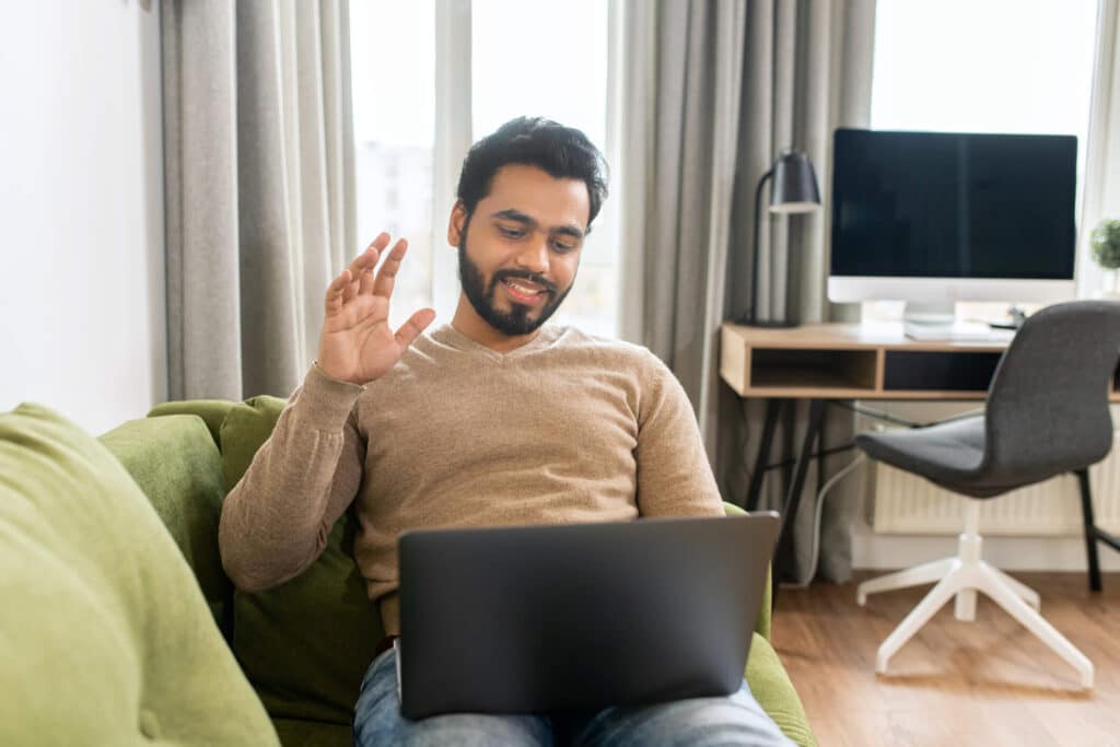 A man sitting on a green sofa with laptop is waving at his therapist in an online consultation