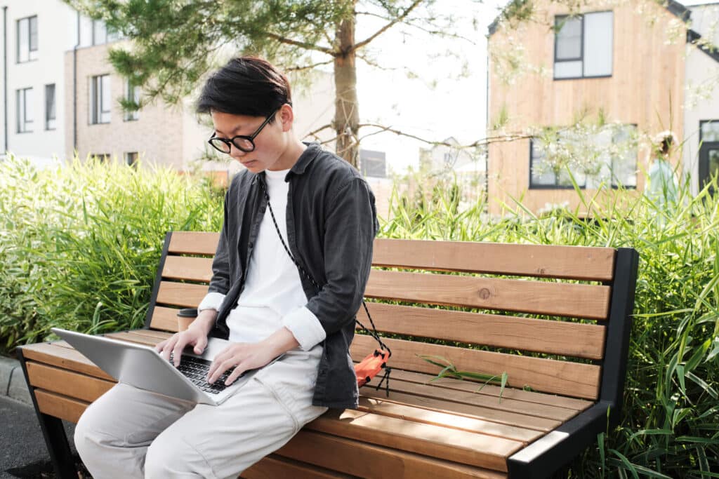 A man wearing glasses sitting on a wooden bench outdoor while working on his laptop