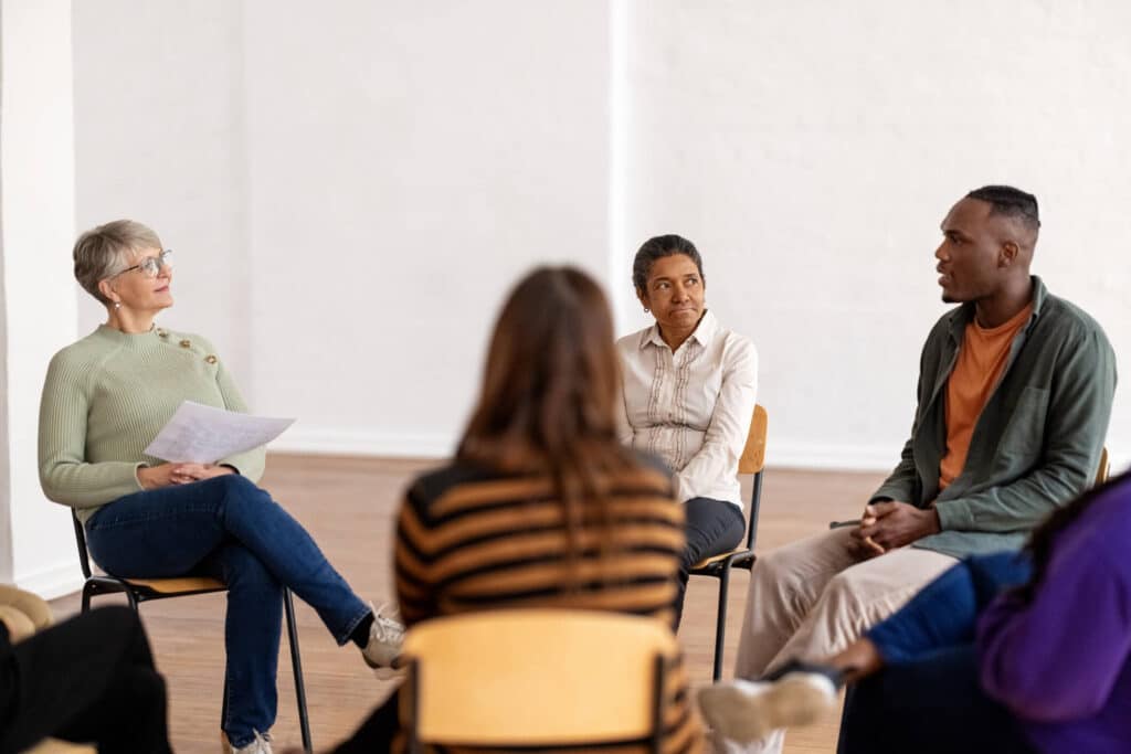 A man talking to a group of people in a group therapy session as they are sitting in a circle, listening and looking at him.