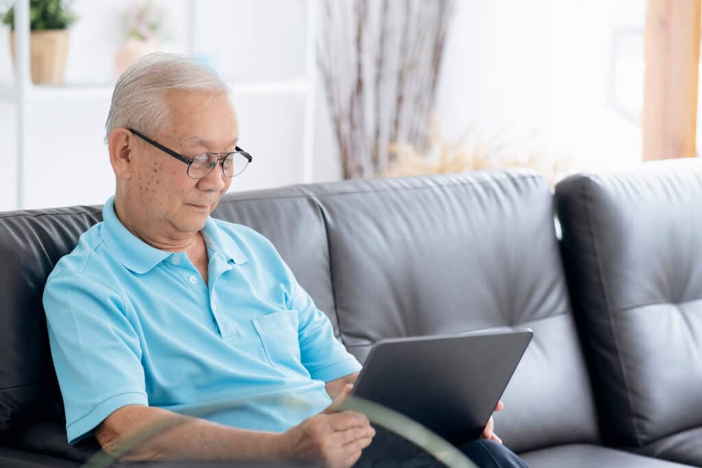 An elderly man wearing glasses comfortably sitting on the sofa  while using an ipad