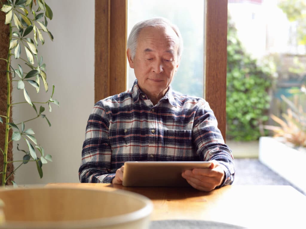 An older gentleman engrossed in using a tablet sitting at a table with a focused expression