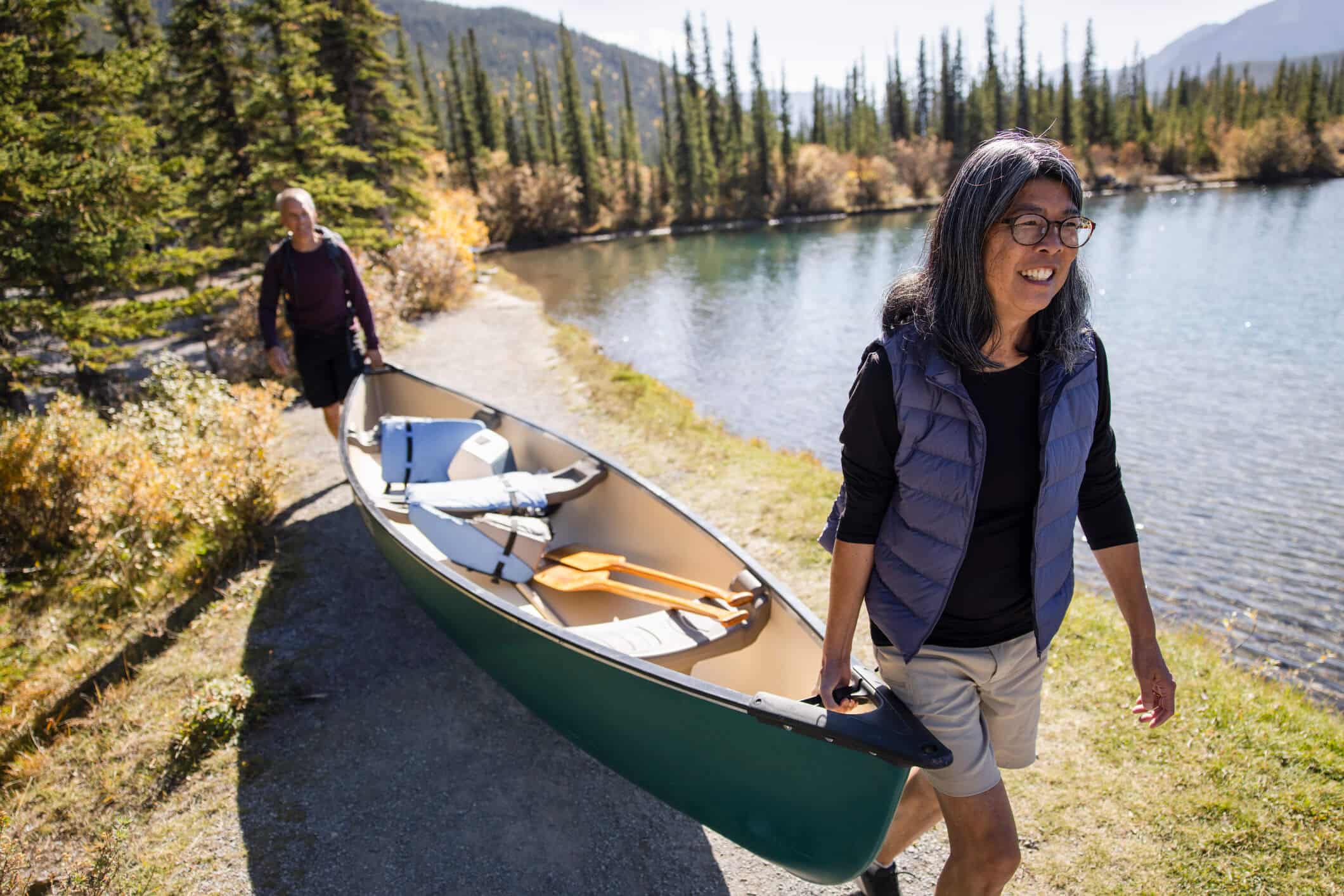 Two happy people walking on a trail by the lake carrying a canoe