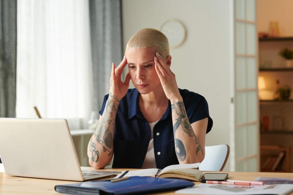 A woman with tattoos on her arms sitting at a desk looking at her a laptop screen while massaging her head
