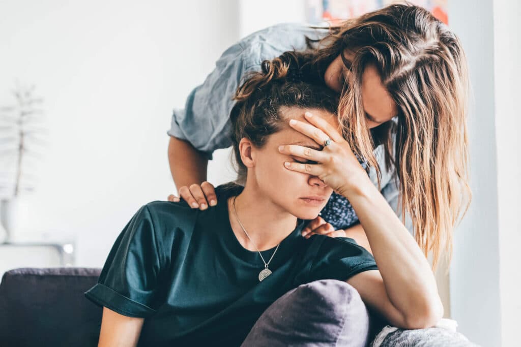Stressed woman holding her head while sitting on a couch and the other woman kissing her head.