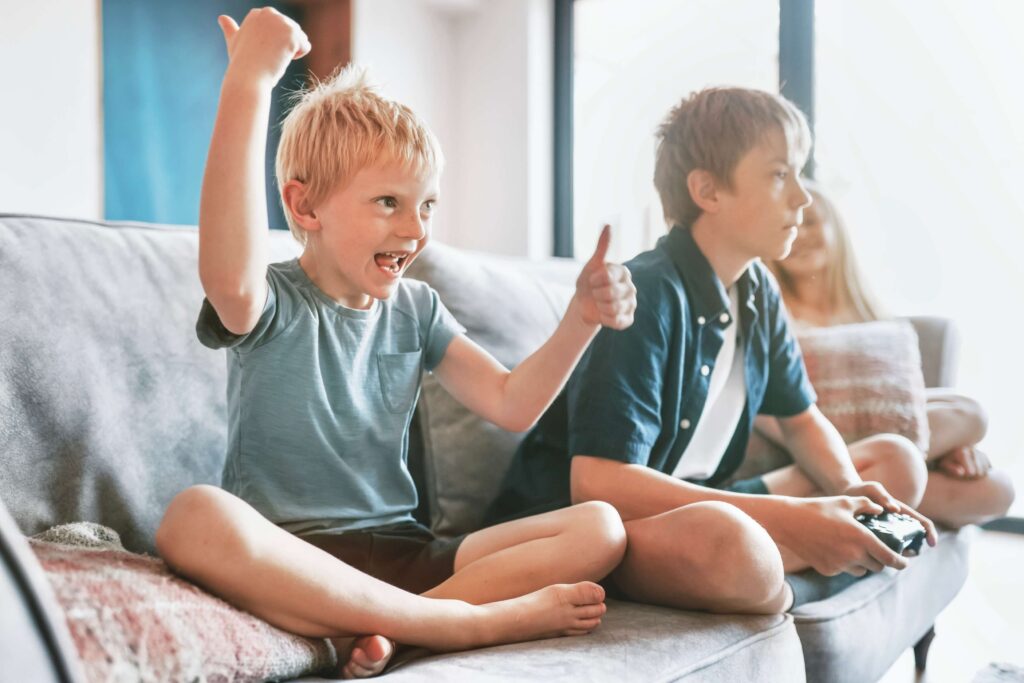 Children happily sitting on a couch one with a big smile on his face while giving thumbs up and the other one playing with a controller