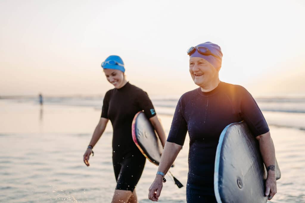 Two smiling women wearing wet suits walking on the beach carrying surfboards