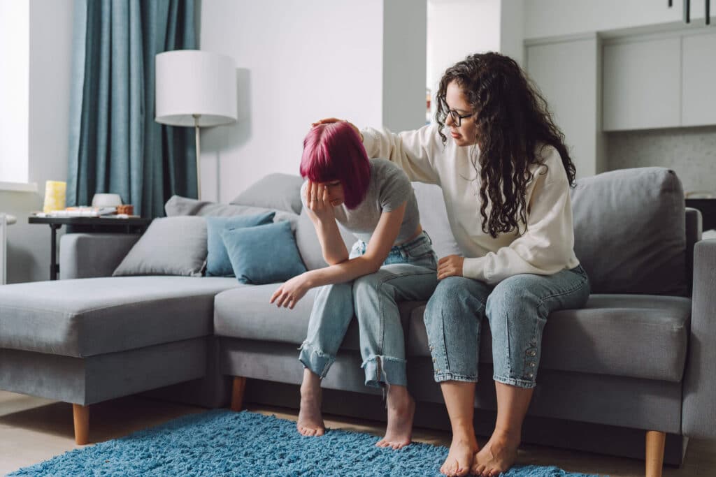 A woman with curly hair comforting the girl with a red hair while both sitting on a couch