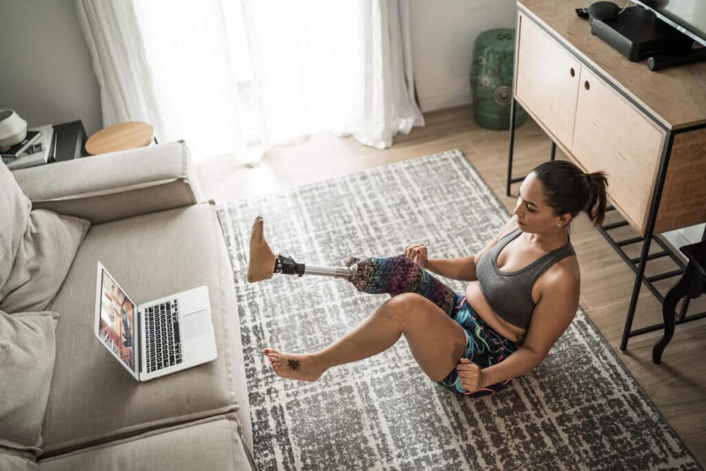 A woman with a prosthetic leg exercising while sitting on the floor in front of her laptop