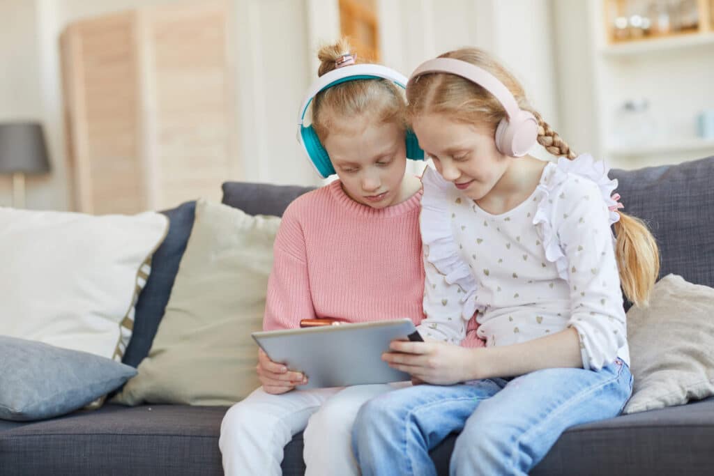 Two young girls wearing heaphones and holding an ipad sitting on a couch while enjoying something from the ipad