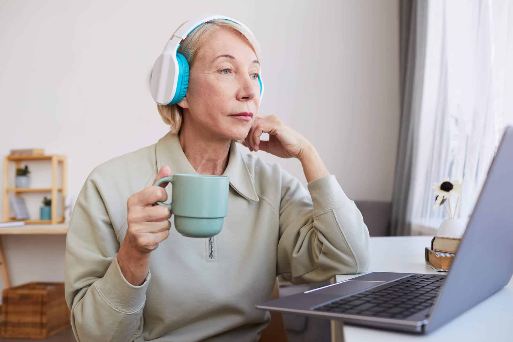 An elderly woman wearing headphones sitting in front of a computer while holding a coffee mug