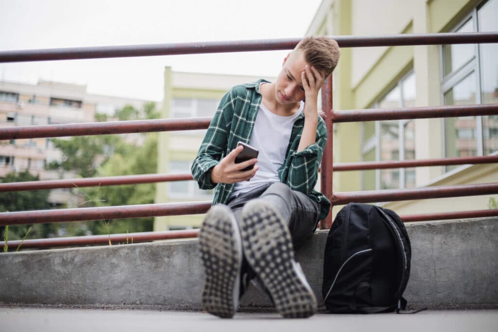A stressed looking boy with a black backpack sits by the railing while looking at his phone