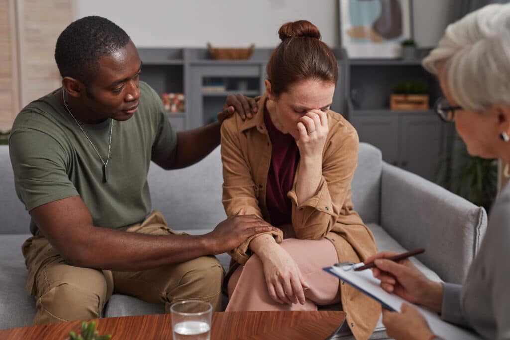 A man consoling a distress looking woman while sitting on a couch during an in person visit with a therapist