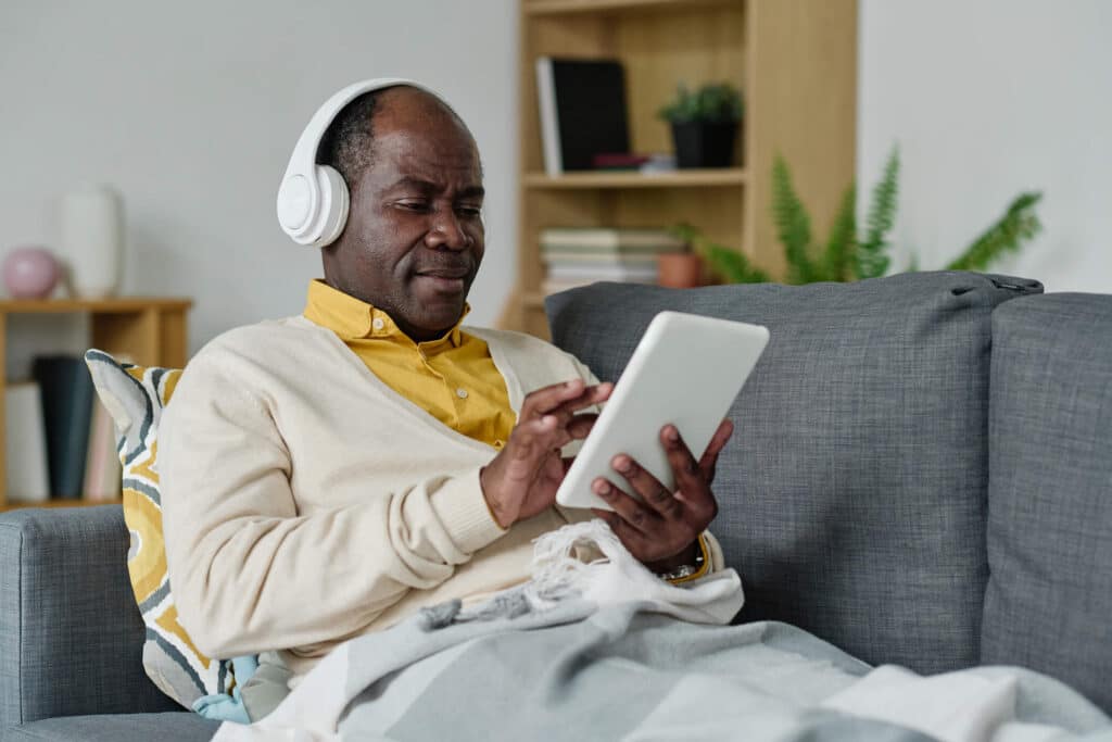 A man wearing headphones sitting comfortably on a couch while watching from his ipad