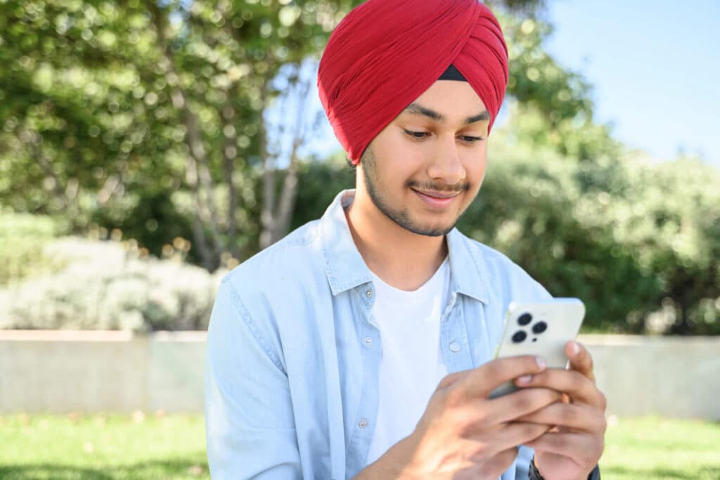 A smiling guy wearing a red turban scrolling through his phone while outdoors