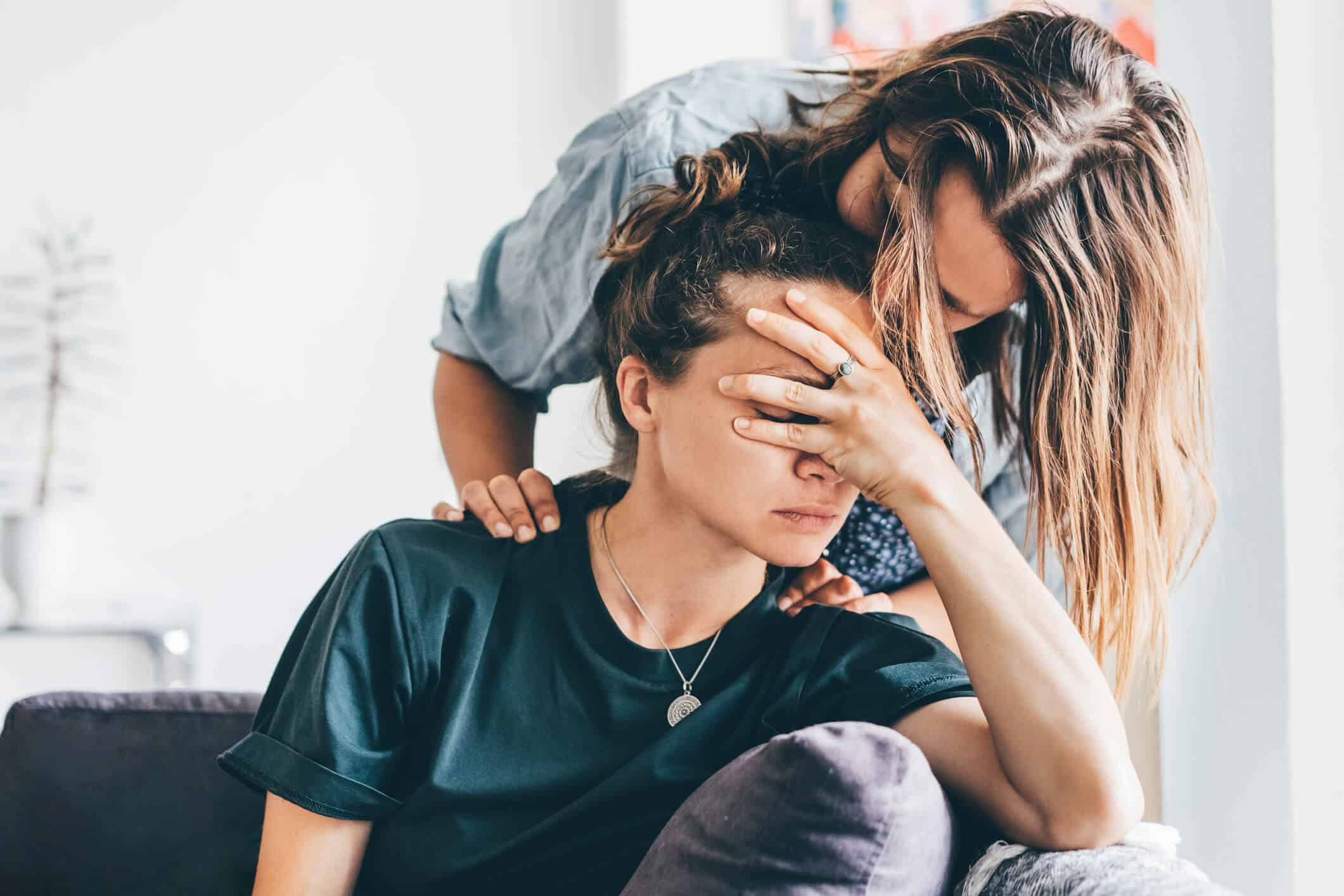 Stressed woman holding her head while sitting on a couch and the other woman kissing her head.