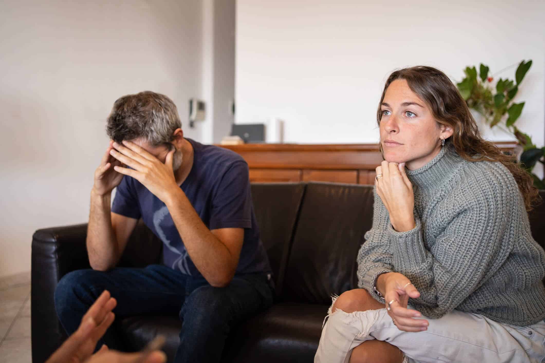 A sad looking man with hands on his head and serious looking lady both sitting on a couch across a therapist during an in person visit