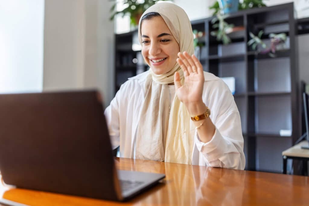 A woman wearing hijab seated at a table in front of a computer waving and smiling on a video call