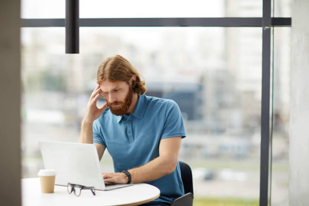 A man wearing blue shirt with his hand on his head seated at a table while working seriously on his laptop