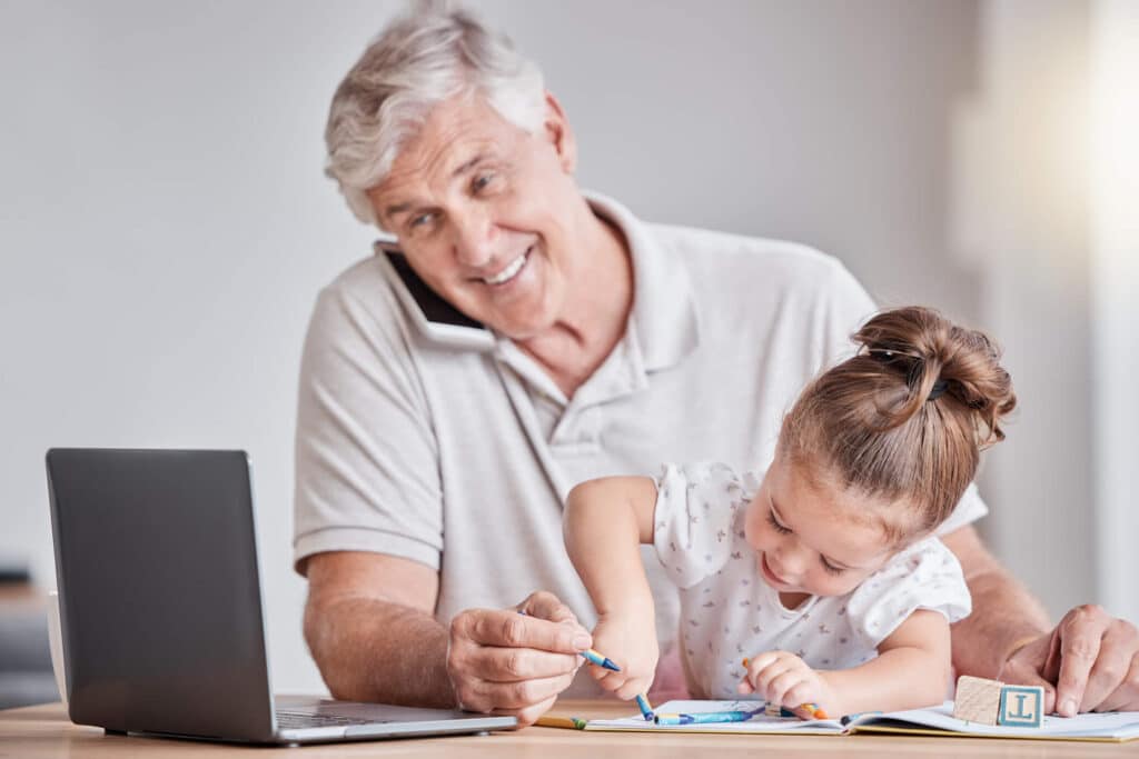 An elderly man talking to someone on the phone while helping the little girl sitting on his lap color the book on the table